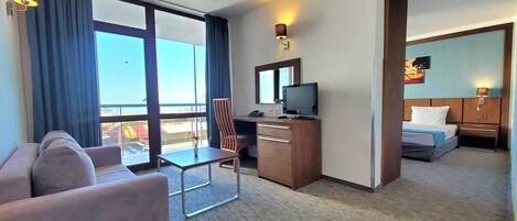 A hotel room with a sofa, a desk with a TV, a chair, a bed, and a large window with curtains.