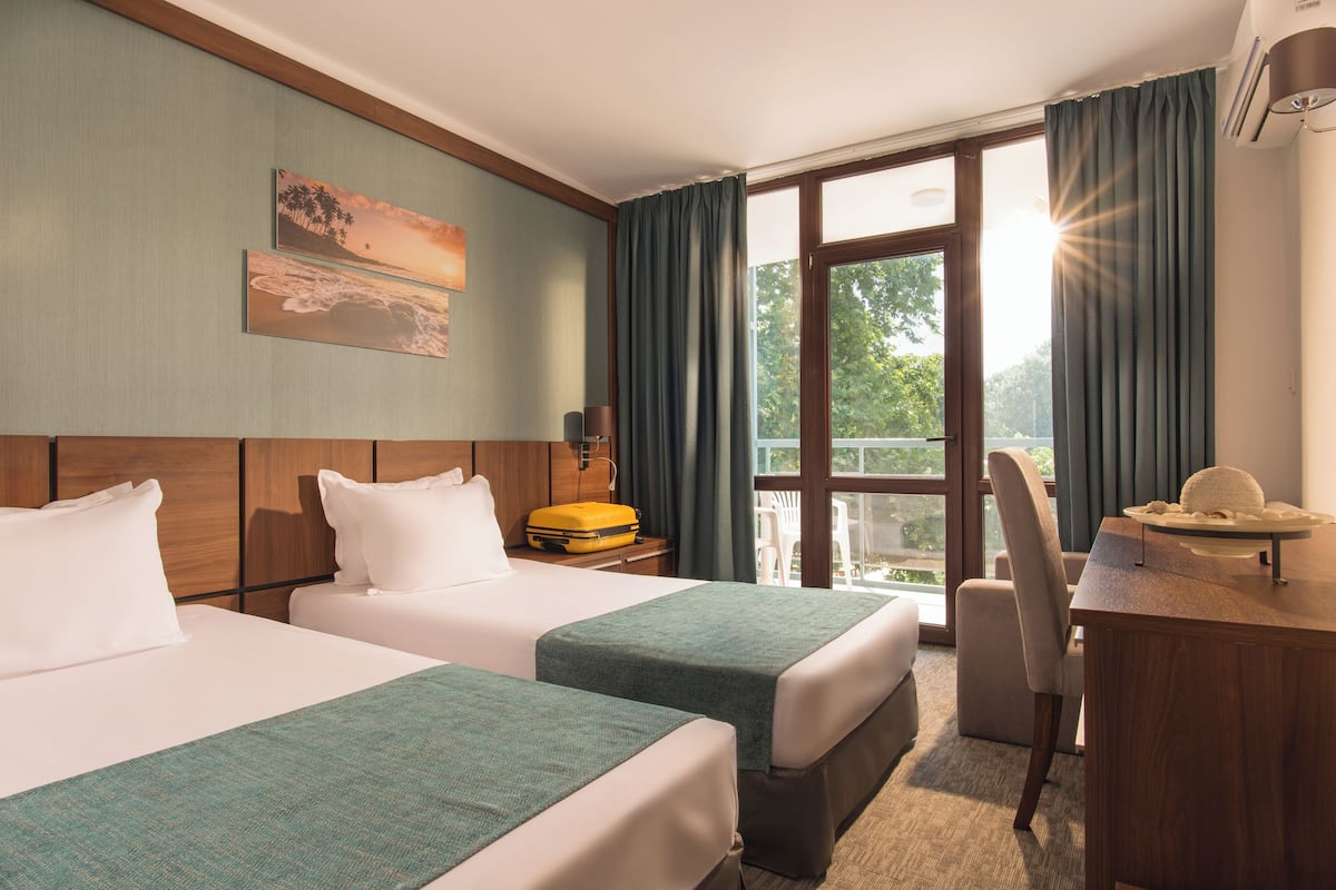 A hotel room with two beds, a desk, a chair, and a large window with curtains.