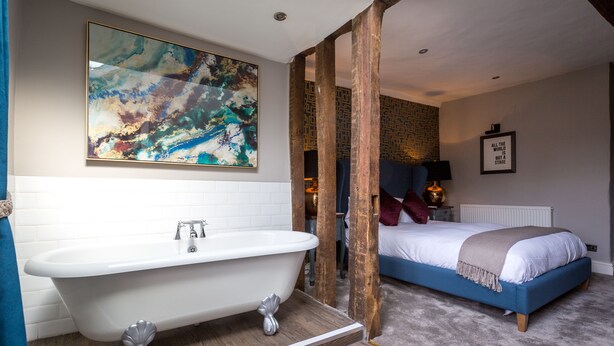 A room with a white bathtub, a large abstract painting, a blue bed, and a wooden floor.