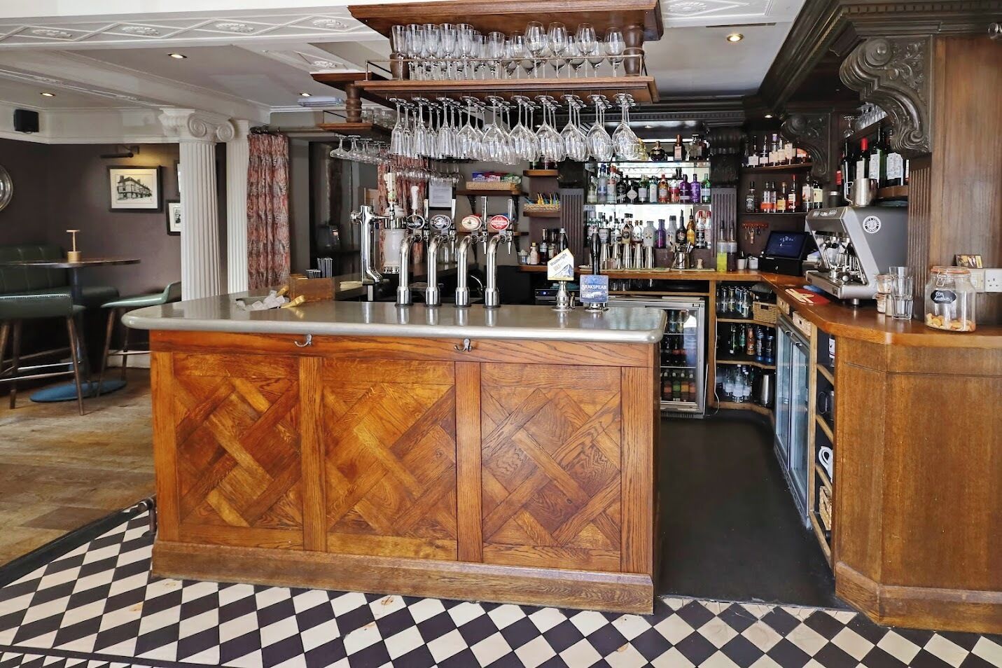 A bar area with a wooden bar counter, multiple taps, and a variety of bottles and glasses.
