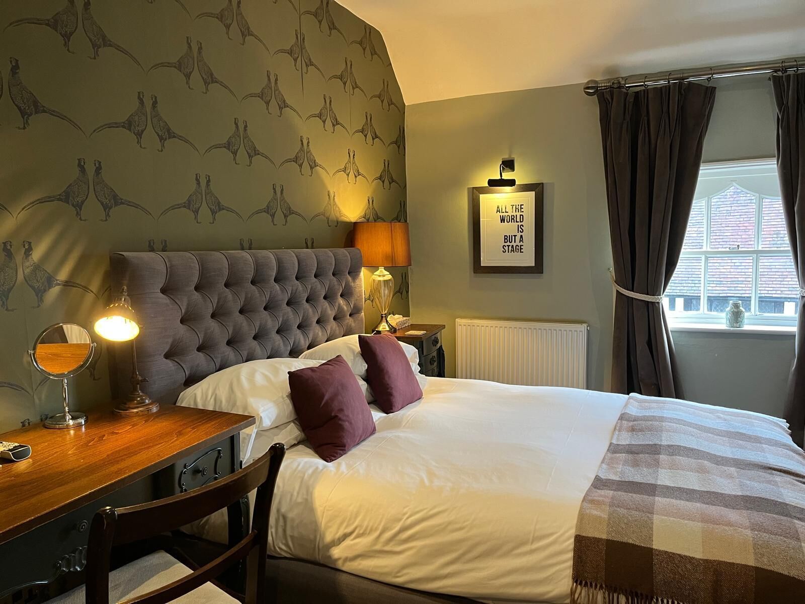 Habitación de hotel con cabecera capitonada, cama con ropa de cama blanca y morada, mesita de noche de madera con lámpara, espejo y una cita enmarcada en la pared.