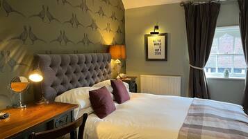 Habitación de hotel con cabecera capitonada, cama con ropa de cama blanca y morada, mesita de noche de madera con lámpara, espejo y una cita enmarcada en la pared.