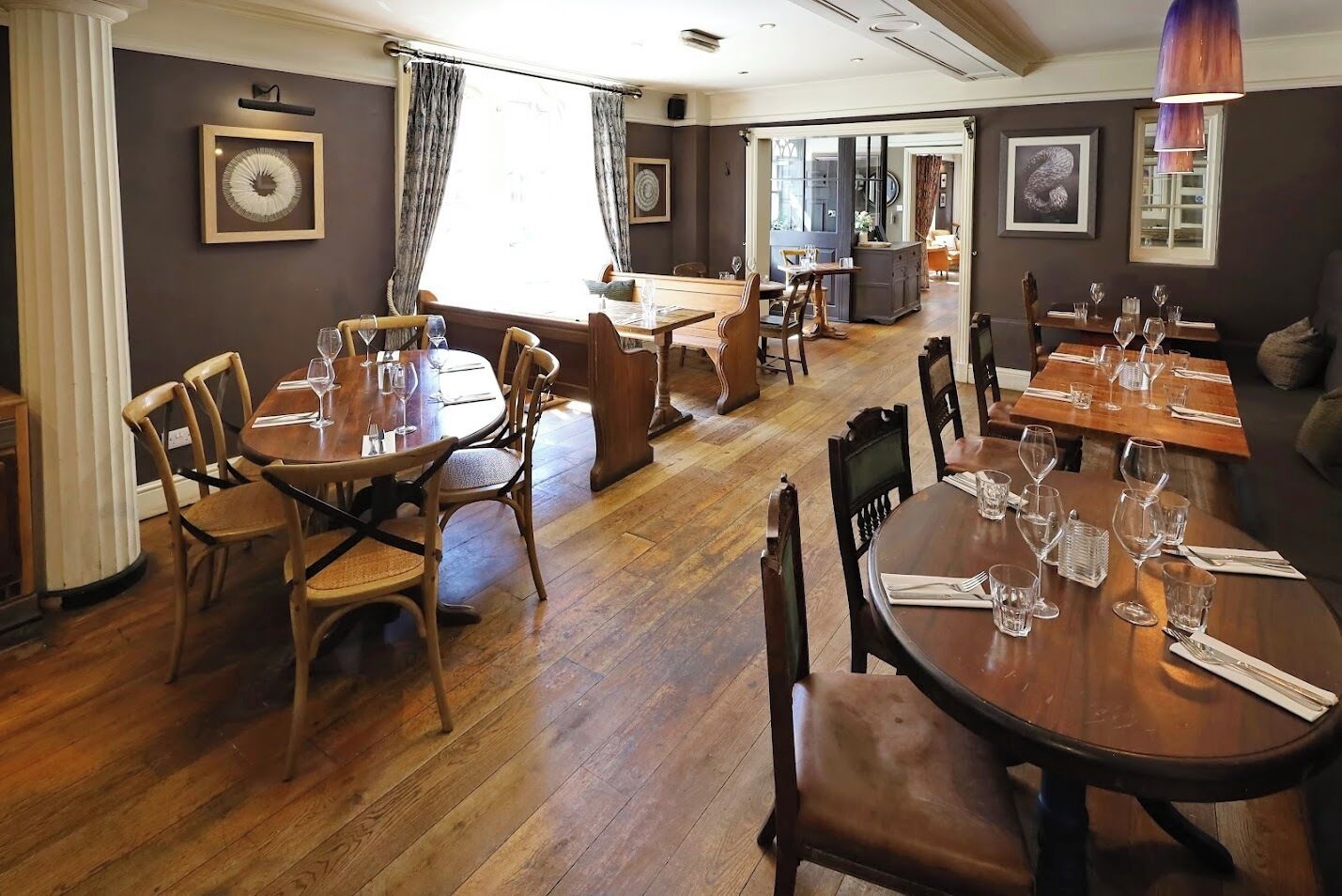 A dining area with wooden tables and chairs, framed artwork on the walls, and a view into another room.