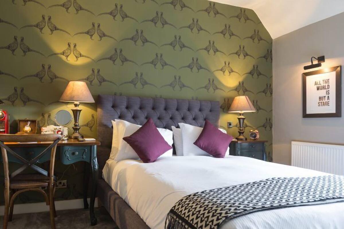 Una habitación de hotel con un cabecero capitonado, una cama con ropa de cama blanca y morada, un escritorio con una lámpara y un papel tapiz con estampado de canguro.