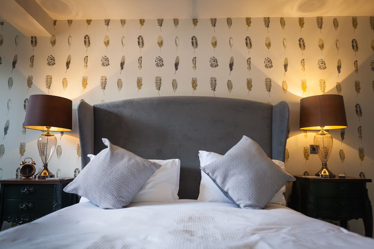 Una cama bien tendida con un cabecero gris, dos almohadas estampadas y dos lámparas de noche a cada lado.