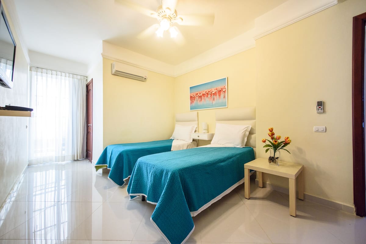 Une chambre d’hôtel avec deux lits, une petite table sur laquelle est posé un vase de fleurs, un ventilateur de plafond et un tableau au mur.