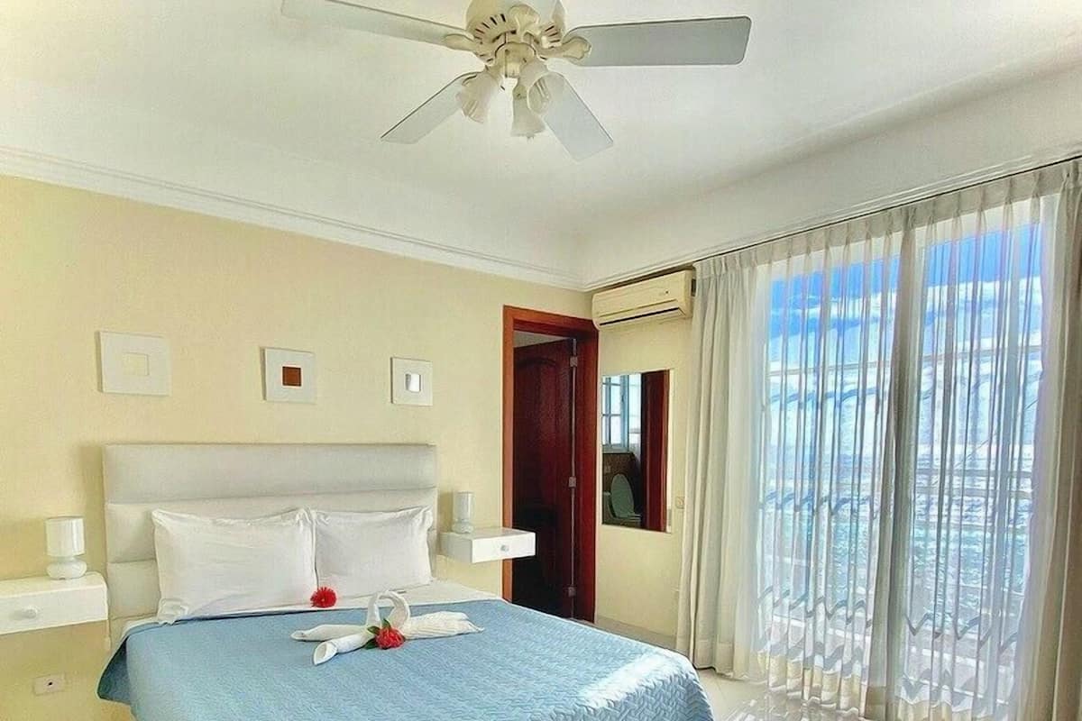 Une chambre d’hôtel avec un lit, un ventilateur de plafond, une table de chevet, une fenêtre avec des rideaux et une vue sur l’extérieur.