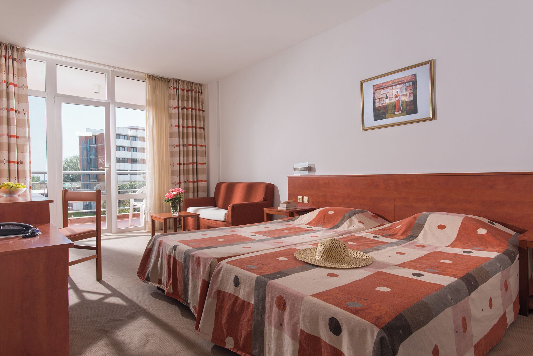A hotel room with a bed, a desk, a chair, a small table with a vase of flowers, and a view through a large window.