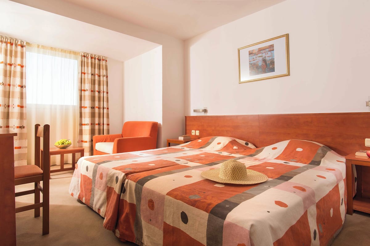 Una habitación de hotel con una cama, una silla, una mesita con un frutero y un cuadro en la pared.
