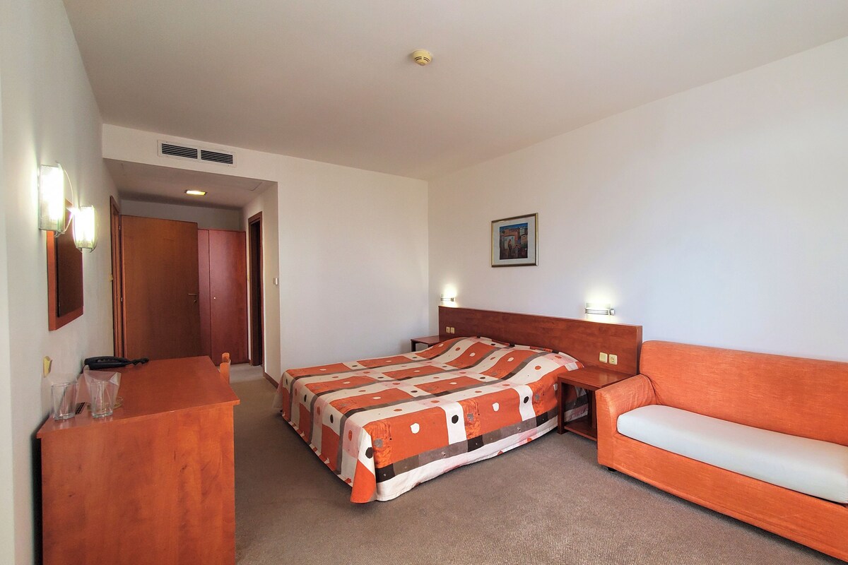 A hotel room with a bed, a wooden dresser, a sofa, and a framed picture on the wall.