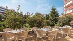 Un patio al aire libre con mobiliario de mimbre y suelo de baldosas, rodeado de vegetación y edificios.
