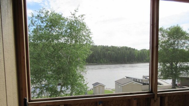 A view through a window with wooden framing, overlooking a river with buildings and trees in the background.