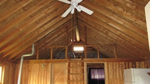 A rustic cabin interior with a wooden ceiling, a ceiling fan, a ladder, a dining table, and a refrigerator.