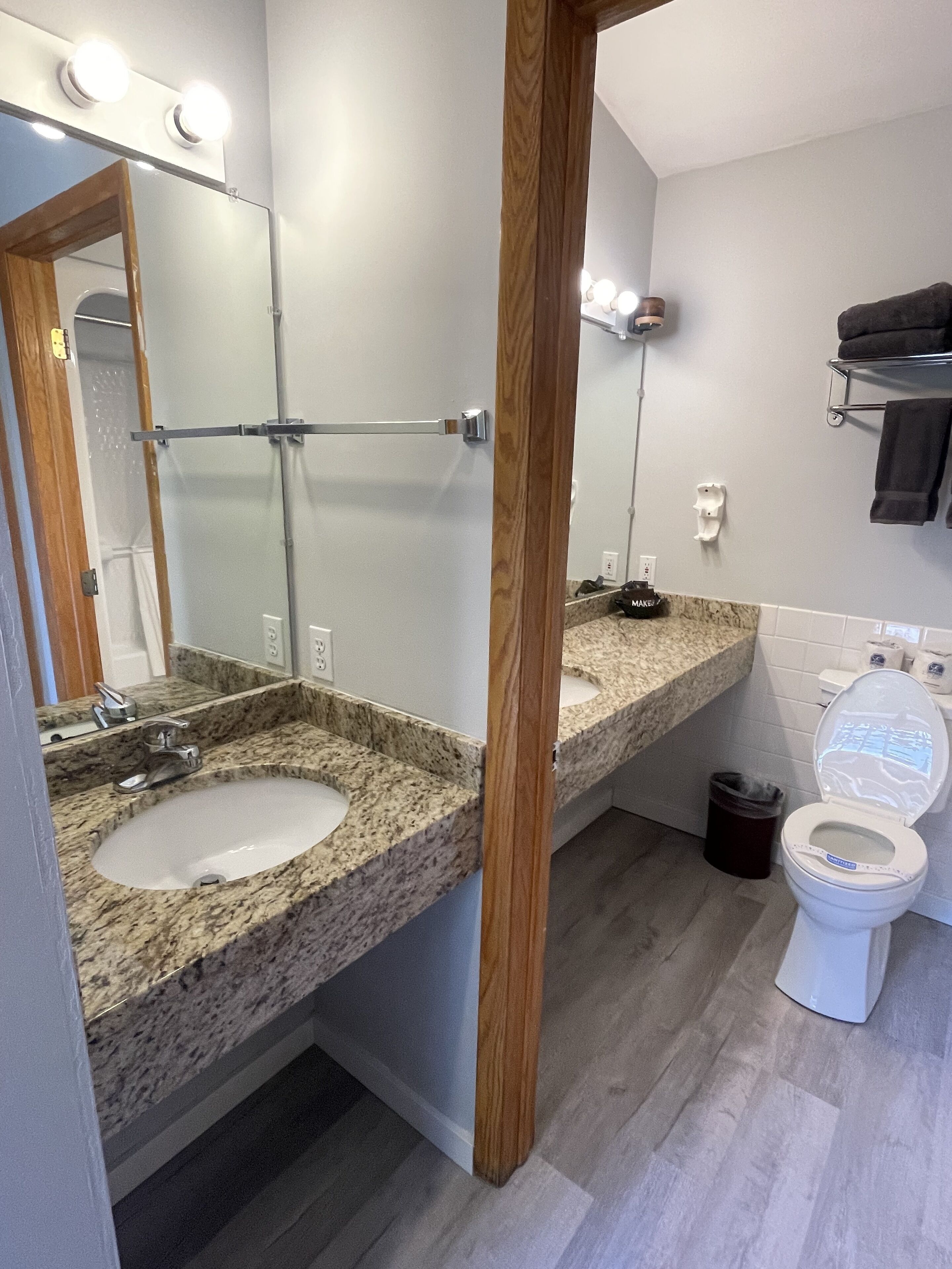 A bathroom with two sinks, a mirror with lights, a toilet, and a trash can.