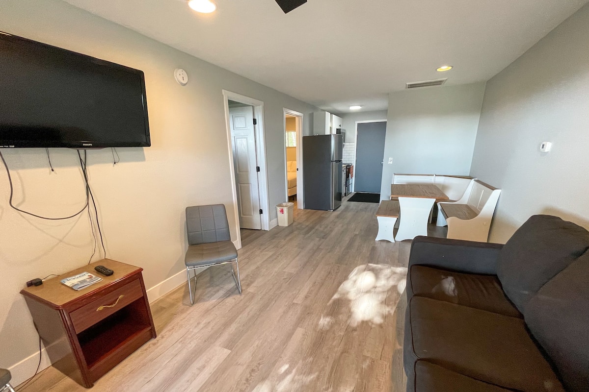 A modern living room with a flat-screen TV, a gray sofa, a wooden coffee table, and a dining area with a white table and chairs.