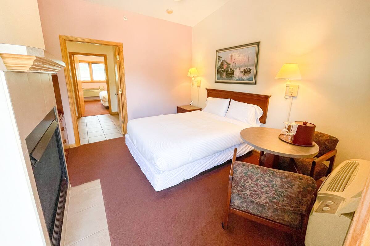 A hotel room with a bed, two bedside lamps, a painting, a small table with a lamp, a chair, and a heater.