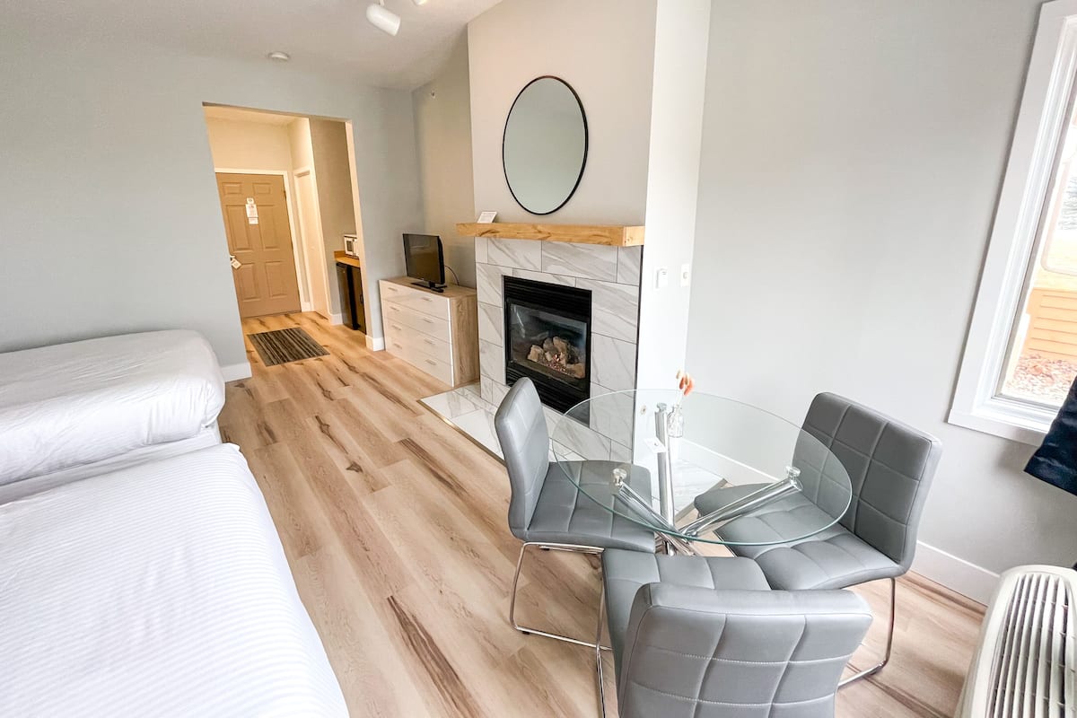 A modern hotel room with a wooden floor, a fireplace, a dining table with chairs, and a large mirror on the wall.