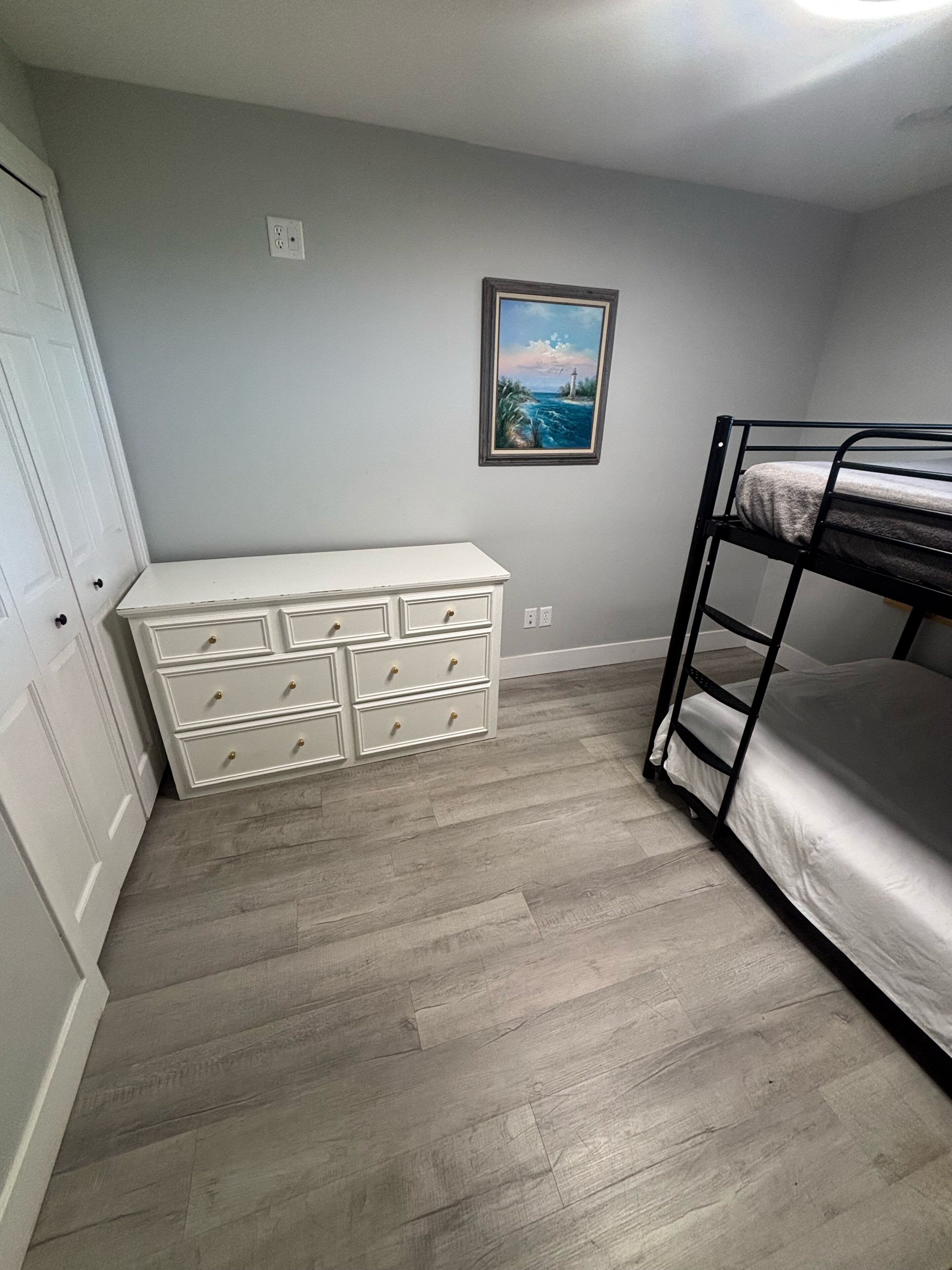 Een kamer met een stapelbed, een witte dressoir met meerdere laden, een lijst met een schilderij aan de muur, en een deur links.