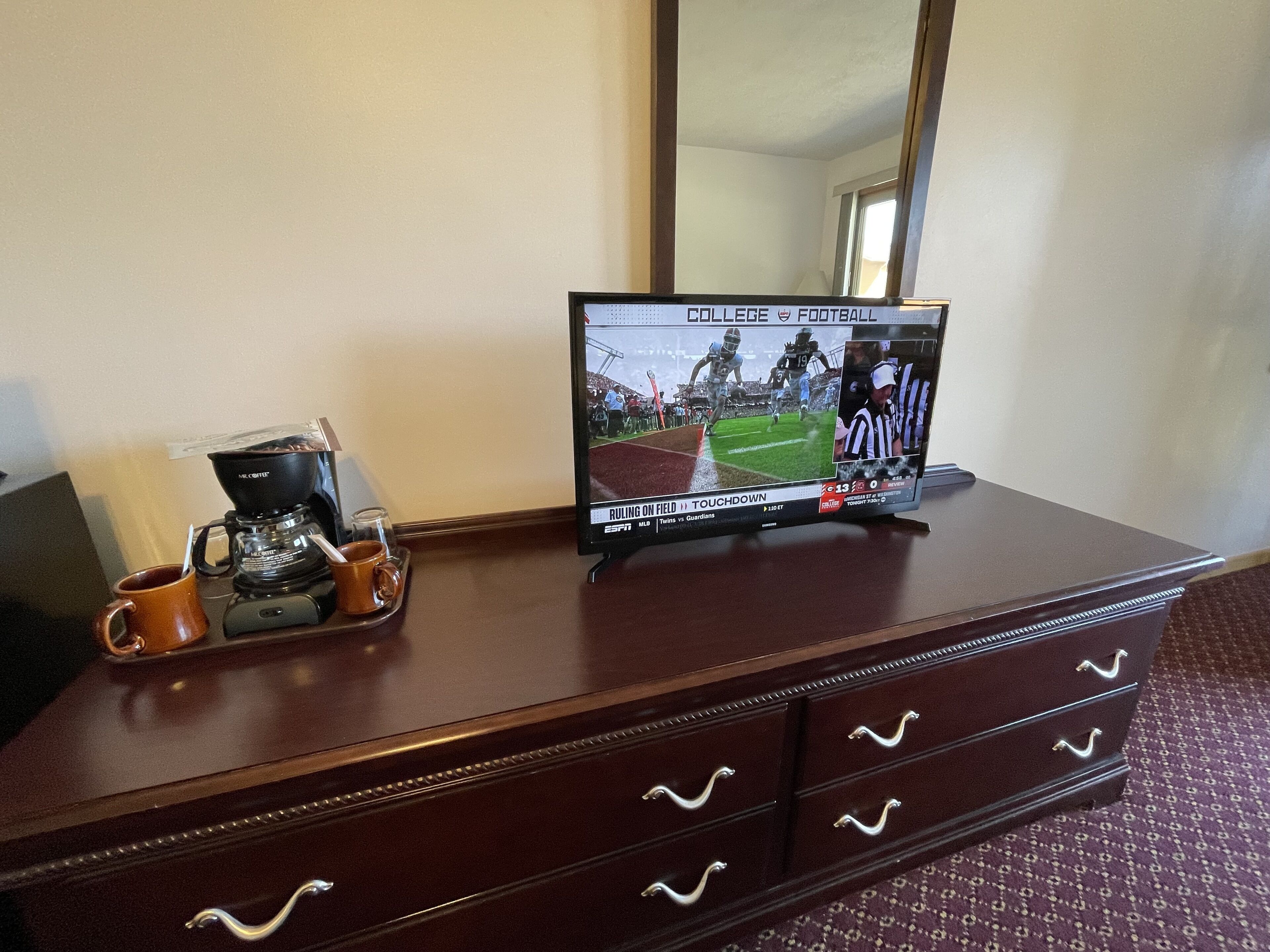 A wooden dresser with a television displaying a football game, a coffee maker, and a mug.