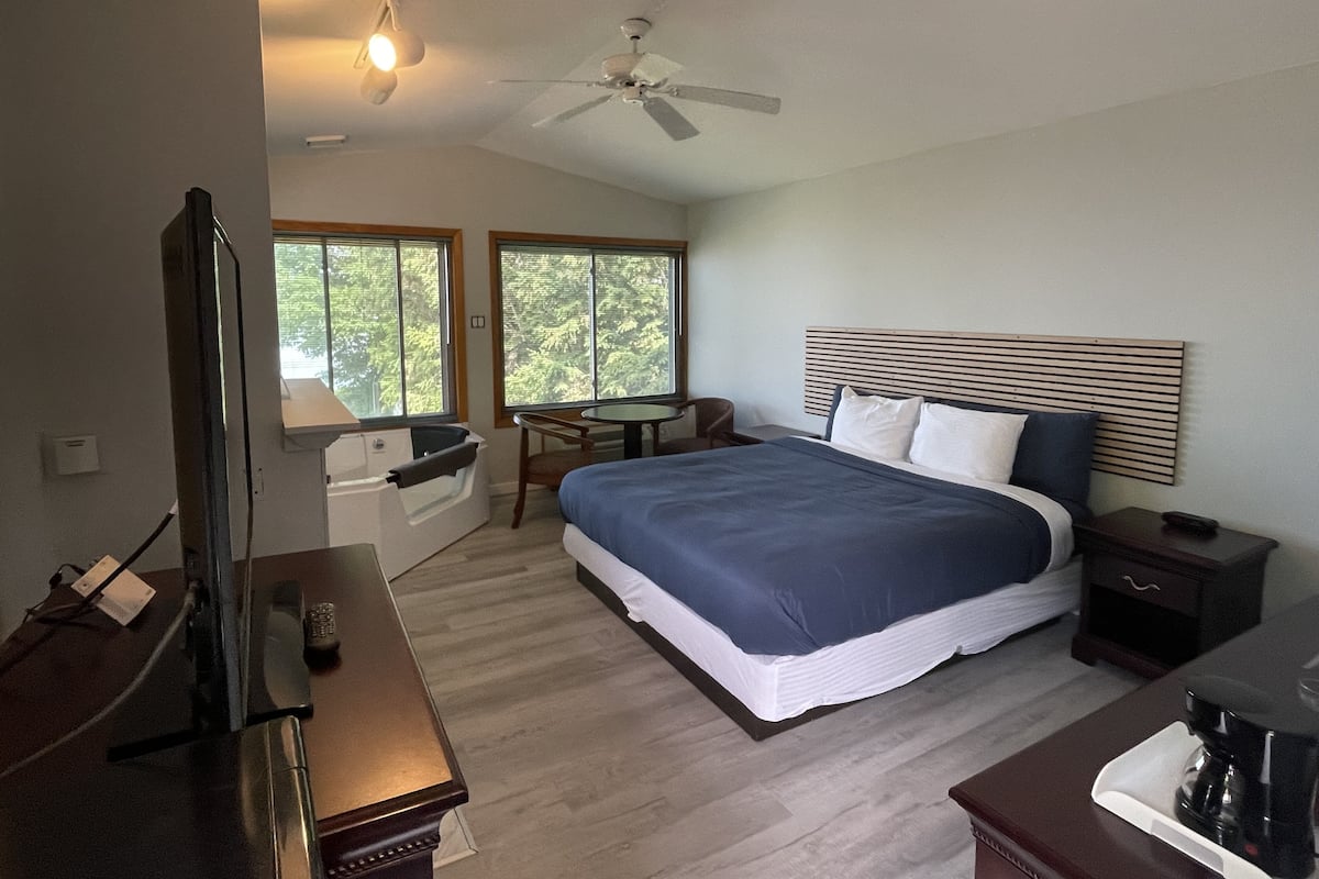 A bedroom with a bed, a desk, a chair, a TV, a ceiling fan, and a window.
