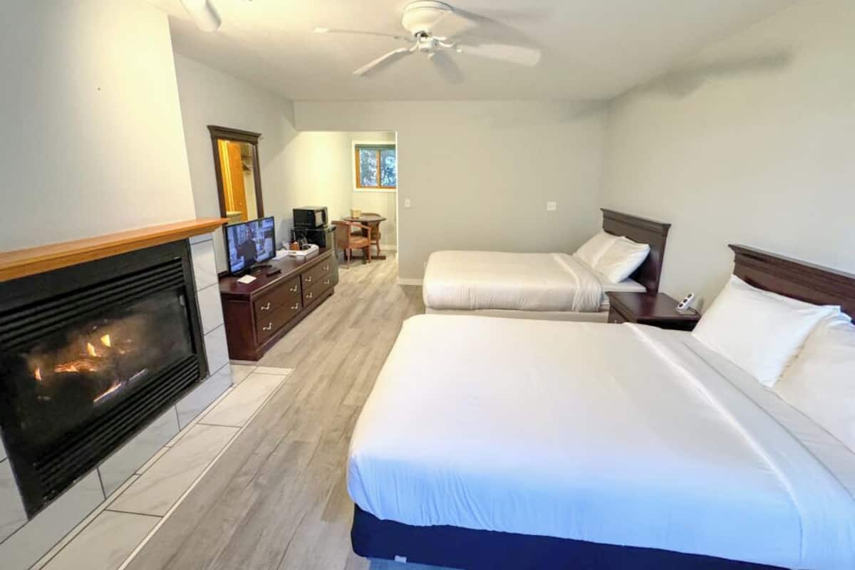 A hotel room with a fireplace, two beds, a desk with a chair, and a ceiling fan.