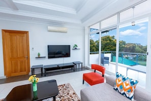 Una sala de estar moderna con un televisor grande, asientos cómodos, una mesa de centro y vistas a un paisaje verde a través de amplios ventanales.