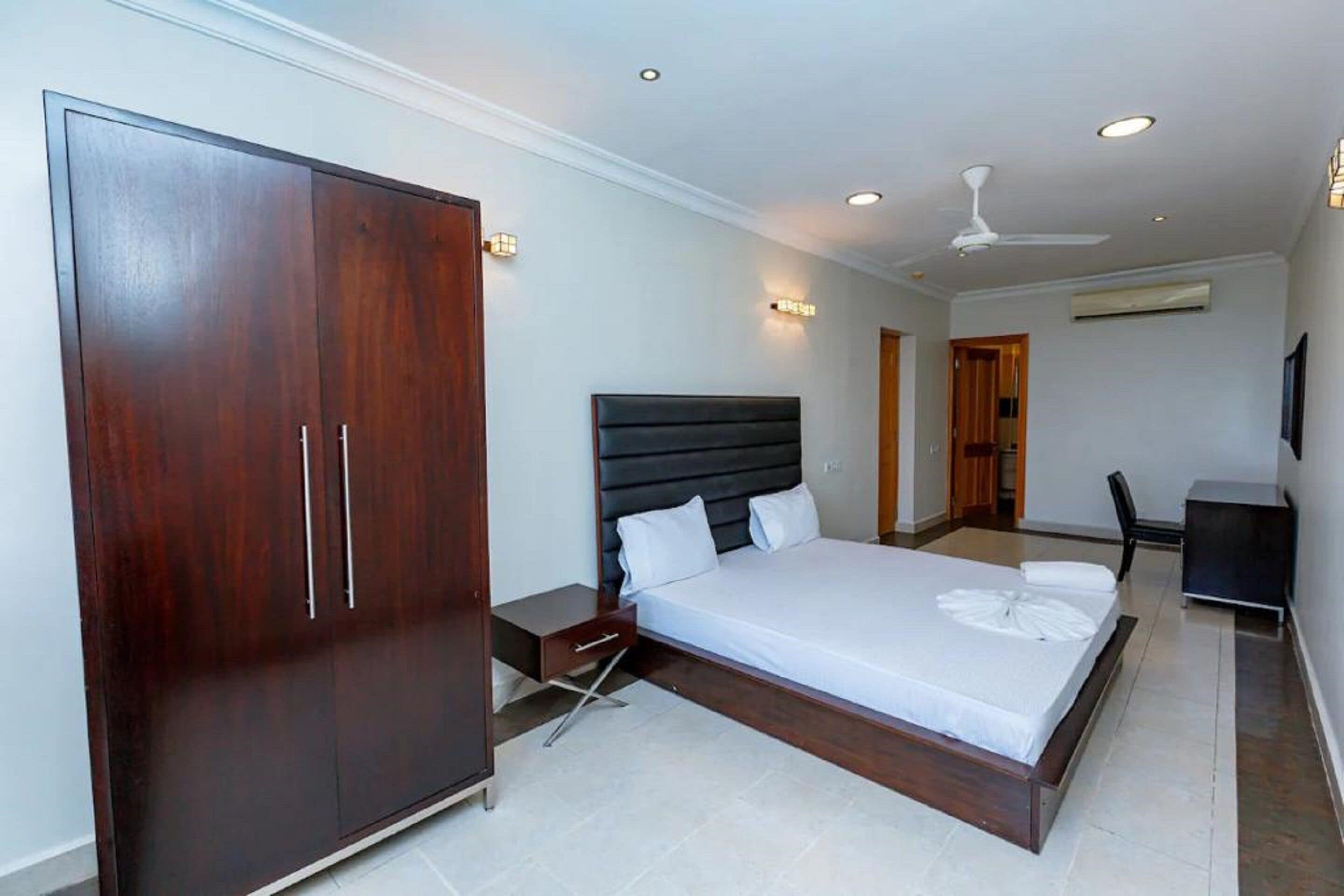 A modern hotel room with a large wardrobe, a bed with white linens, a desk, and a ceiling fan.