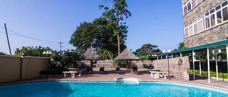 A swimming pool with lounge chairs and thatched umbrellas, surrounded by a stone building and trees.