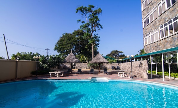 Piscina con sillones de descanso y sombrillas de paja, rodeada de un edificio de piedra y árboles.