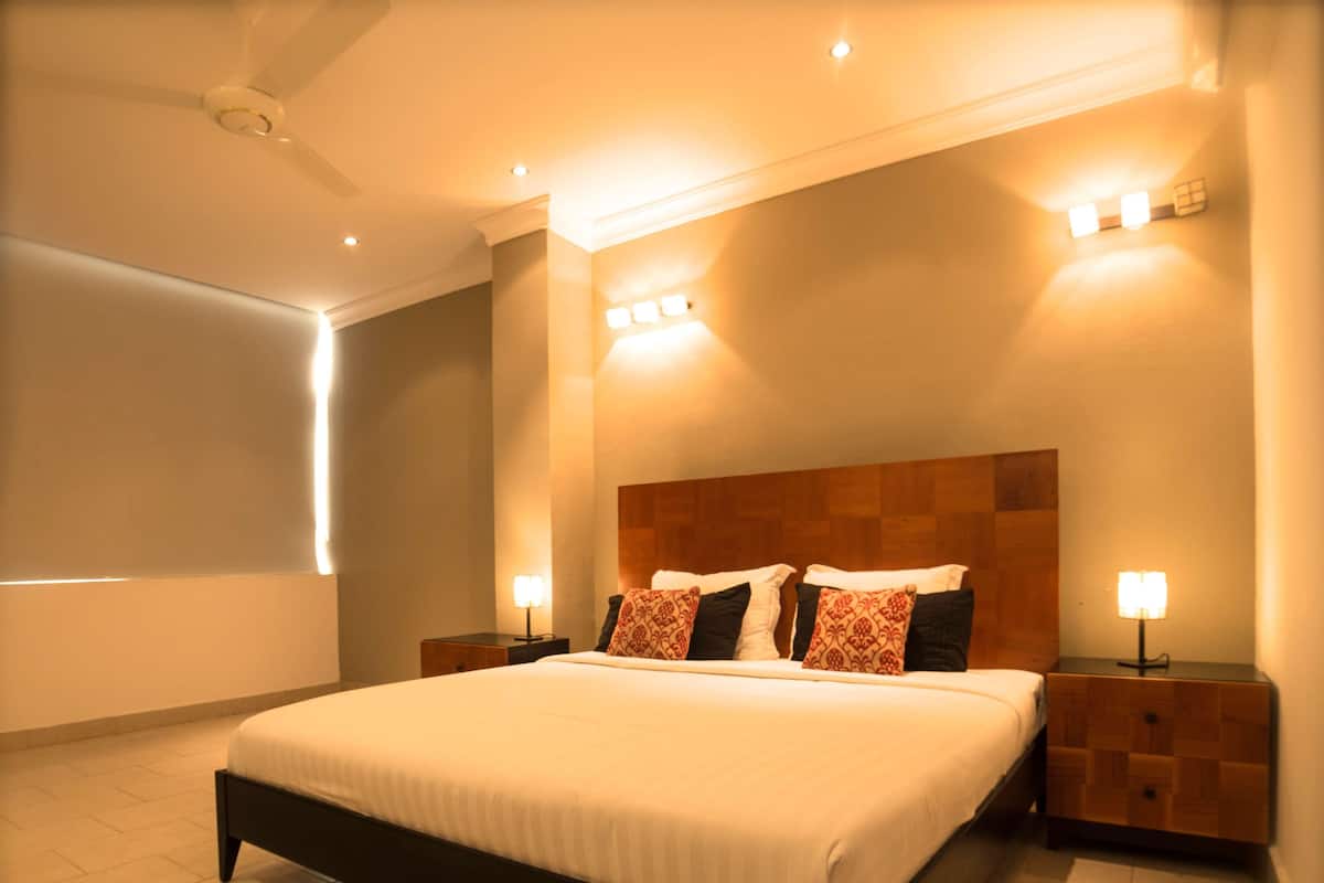 Une chambre à coucher moderne avec un grand lit, une tête de lit en bois, deux tables de chevet avec des lampes et un luminaire au plafond.
