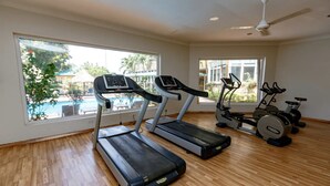 Een sportschool met loopbanden, een elliptische machine en een hometrainer. Grote ramen bieden uitzicht op een zwembad en groen.