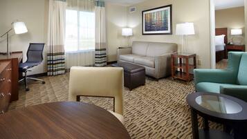 A hotel room with a sofa, armchair, ottoman, a round table, a desk with a lamp, a chair, and a bed in the background.
