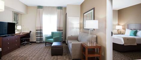 A hotel room with a desk, chair, sofa, ottoman, bedside table, lamp, and a bed.
