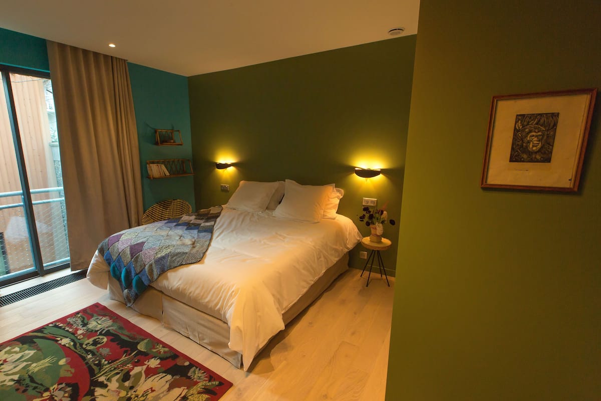 A bedroom with a large bed, a window with curtains, a small bedside table with a vase, and a framed picture on the wall.