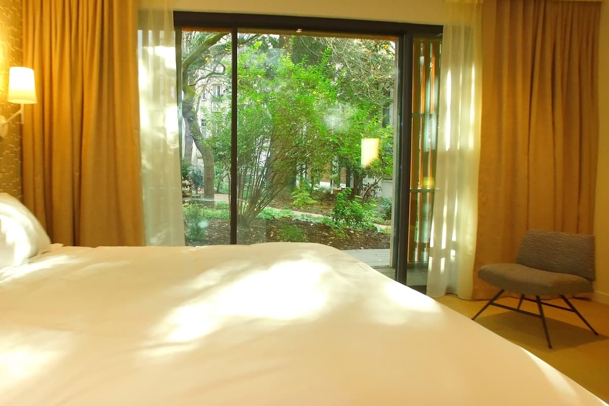 A hotel room with a large bed, a chair, a window with curtains, and a wall-mounted lamp.