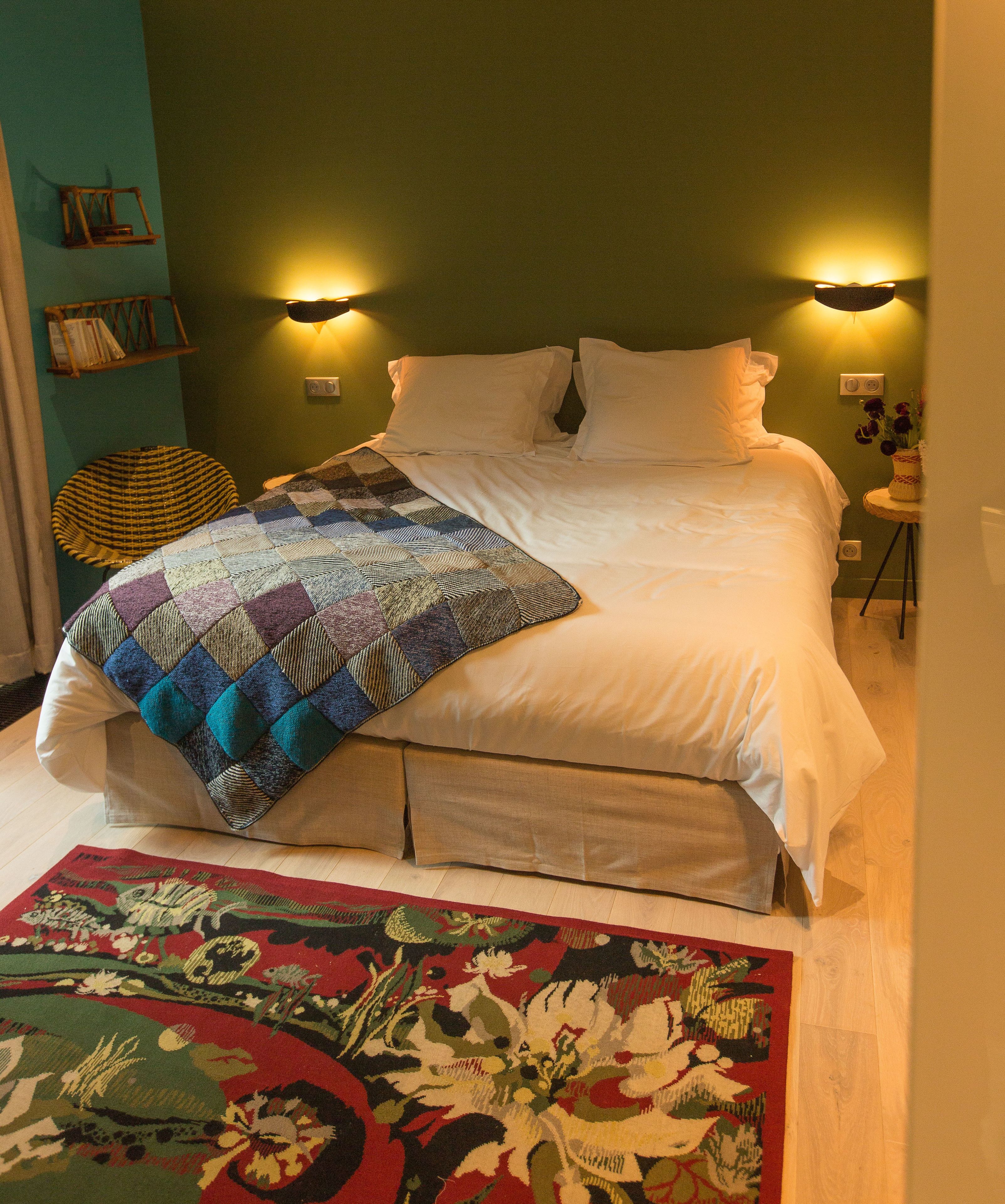 Un lit bien fait, recouvert d’une courtepointe, une petite table ronde avec un vase, et une chaise dont l’assise est en osier.