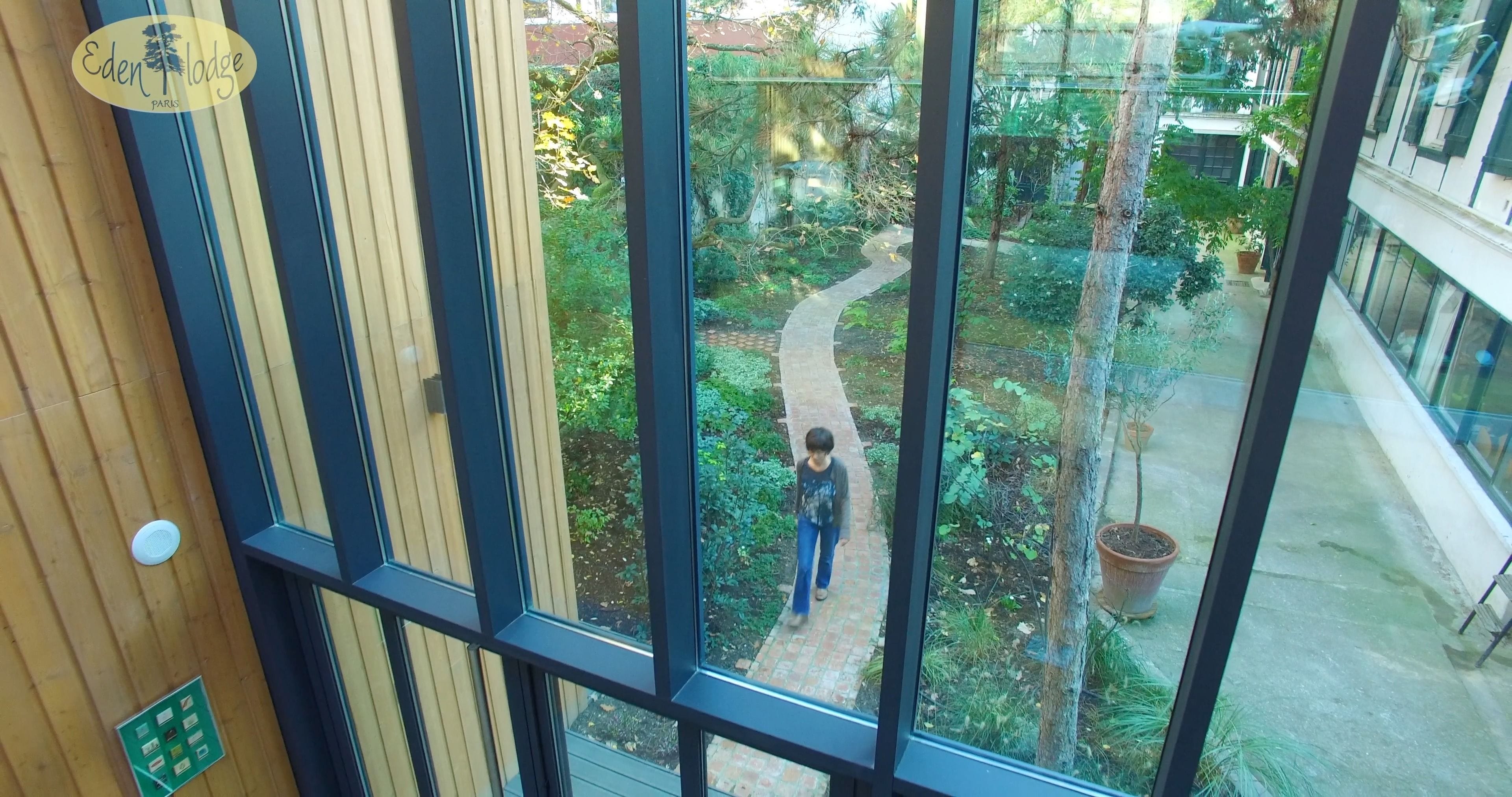 Blick aus einem Fenster eines Gebäudes mit Holzinnenraum und Glaswand auf einen Gartenpfad mit viel Grün.