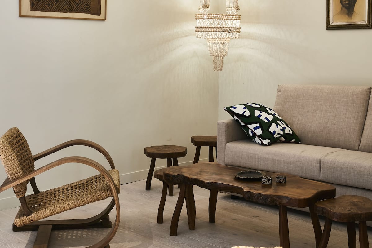 A living room with a rattan chair, wooden coffee table, and a sofa with a patterned pillow. A chandelier hangs from the ceiling, and artwork is displayed on the walls.