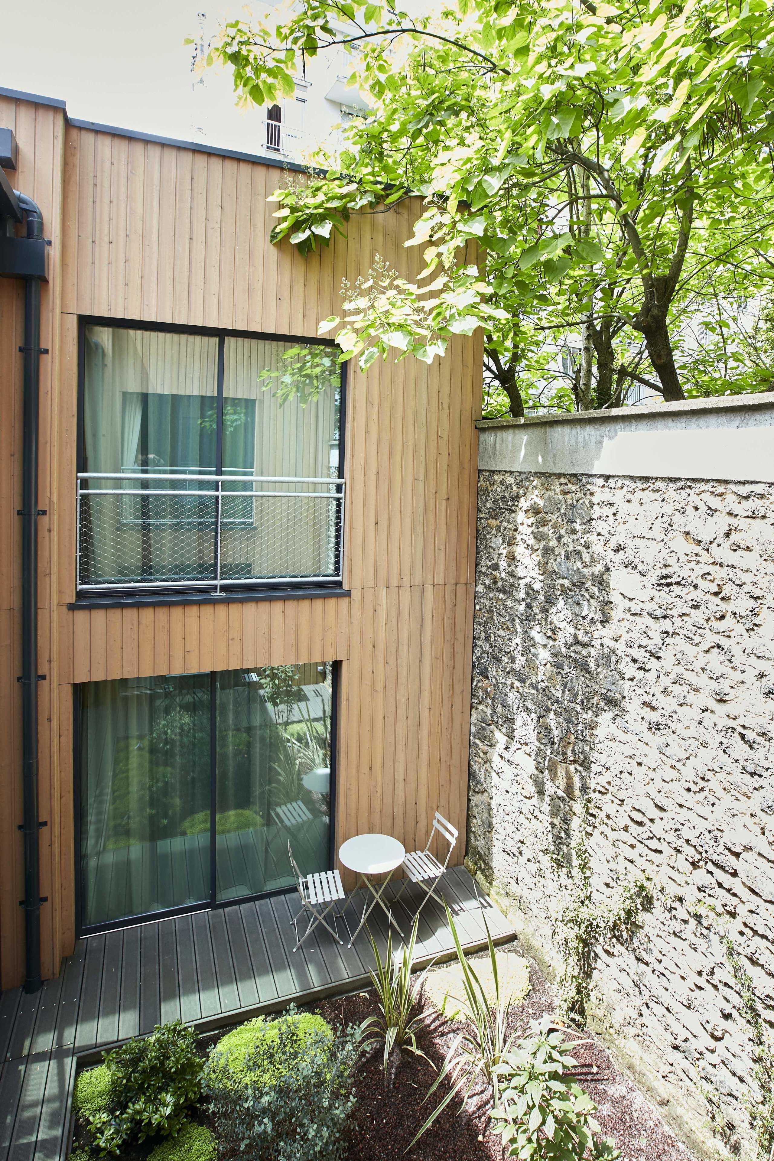 A modern building with a wooden exterior, large windows, and a small outdoor seating area with a table and chairs.