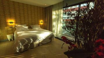 A modern hotel room with a large bed, two bedside tables, and a view of a building through sheer curtains.