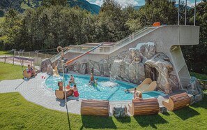 A swimming pool area with a slide, wooden benches, and a rocky backdrop.