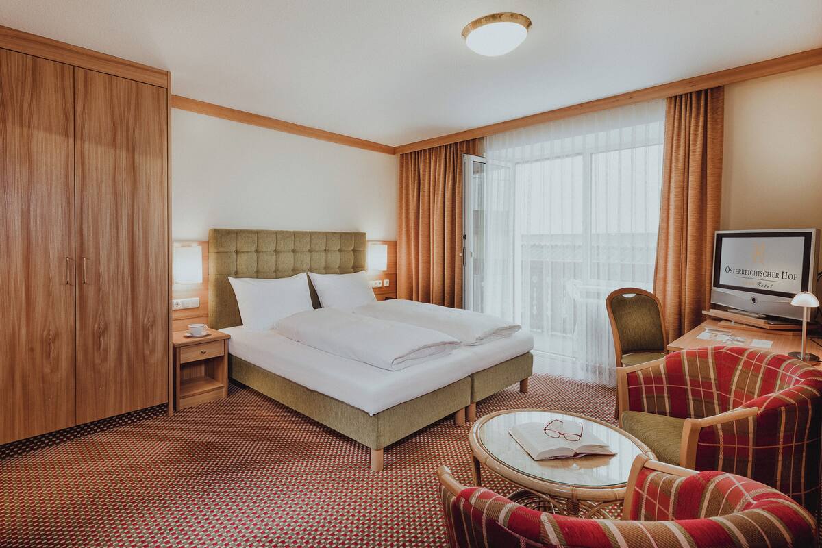 Ein Hotelzimmer mit einem Bett, einem Schreibtisch mit Computer, einem Sessel und einem kleinen Tisch mit Buch und Gläsern.