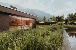 A modern building with large glass windows, a wooden deck, and a view of mountains. - Kurhotel Österreichischer Hof (Bad Hofgastein)