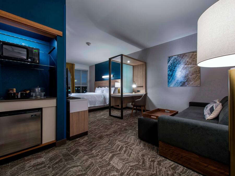 A hotel room with a kitchenette, a bed, a desk, a sofa, and a lamp.