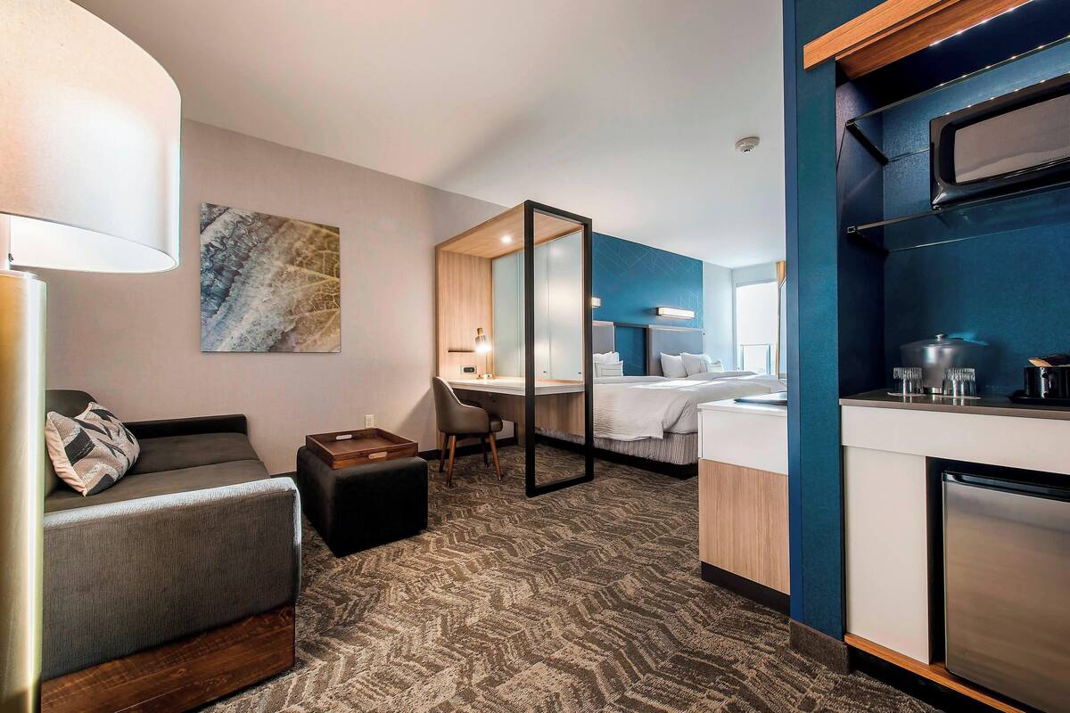 A hotel room with a sofa, a desk, a bed, a kitchenette, and a large mirror.