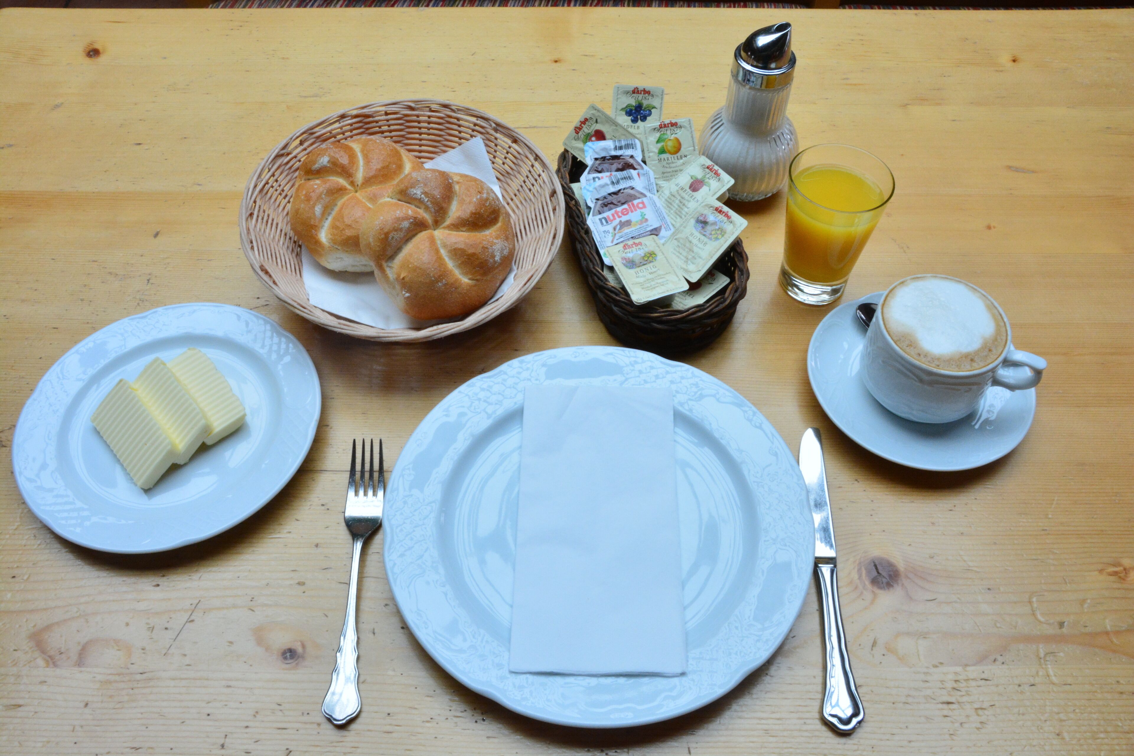 A breakfast setting with bread, butter, a basket, a glass of orange juice, and a cup of coffee on a wooden table.