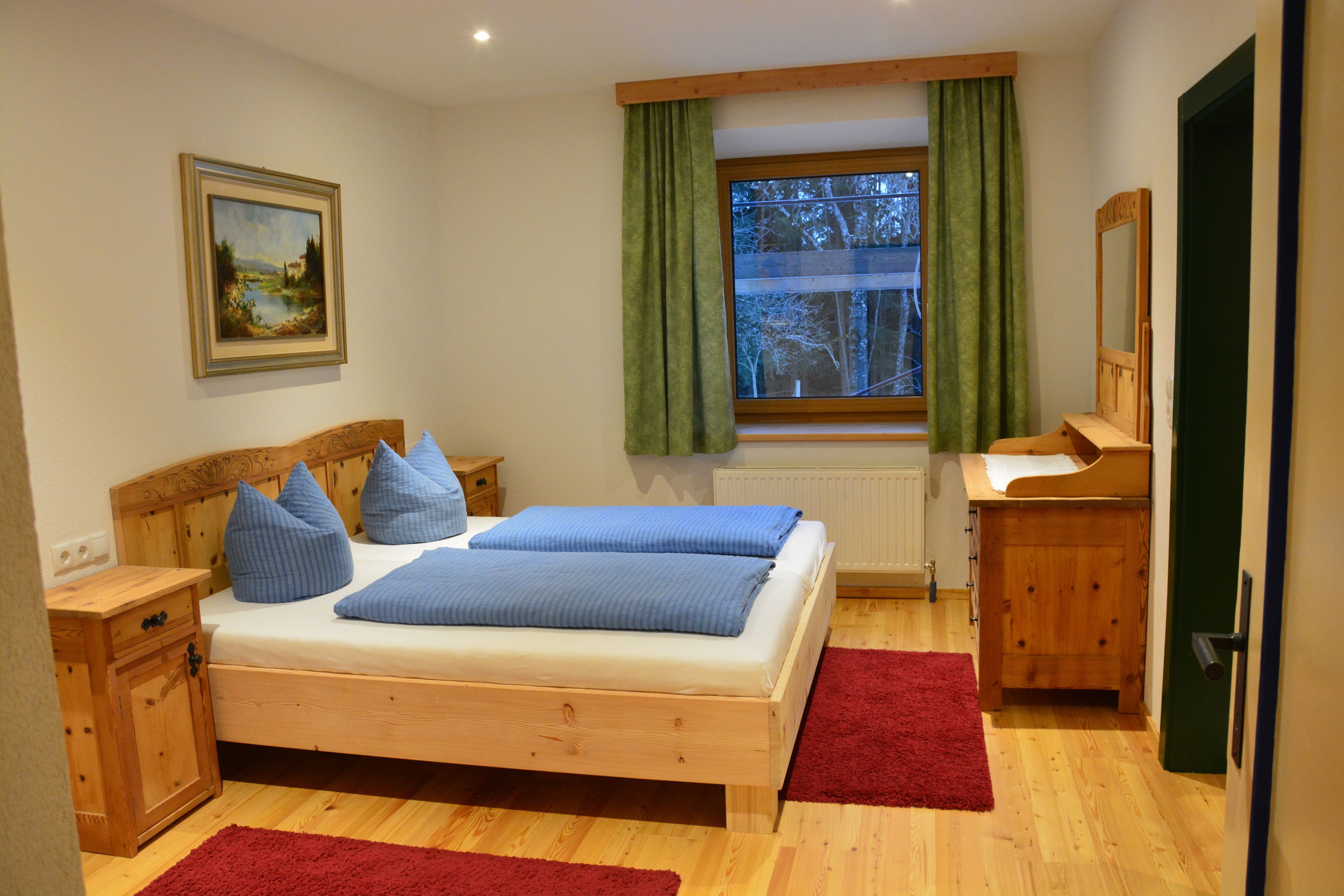 Une chambre à coucher avec un lit en bois, une table de chevet, une commode, une fenêtre avec des rideaux et un tableau accroché au mur.