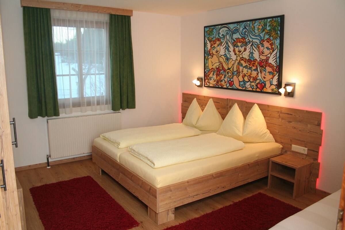 A double bed with a wooden headboard, two bedside tables, a red rug, a window with green curtains, and a colorful painting on the wall.