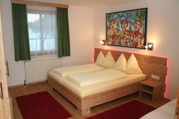 A double bed with a wooden headboard, two bedside tables, a red rug, a window with green curtains, and a colorful painting on the wall.