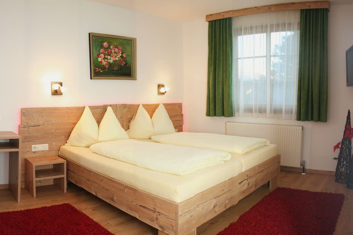 A double bed with a wooden headboard, two bedside tables, a red rug, a green curtain, and a painting of flowers on the wall.